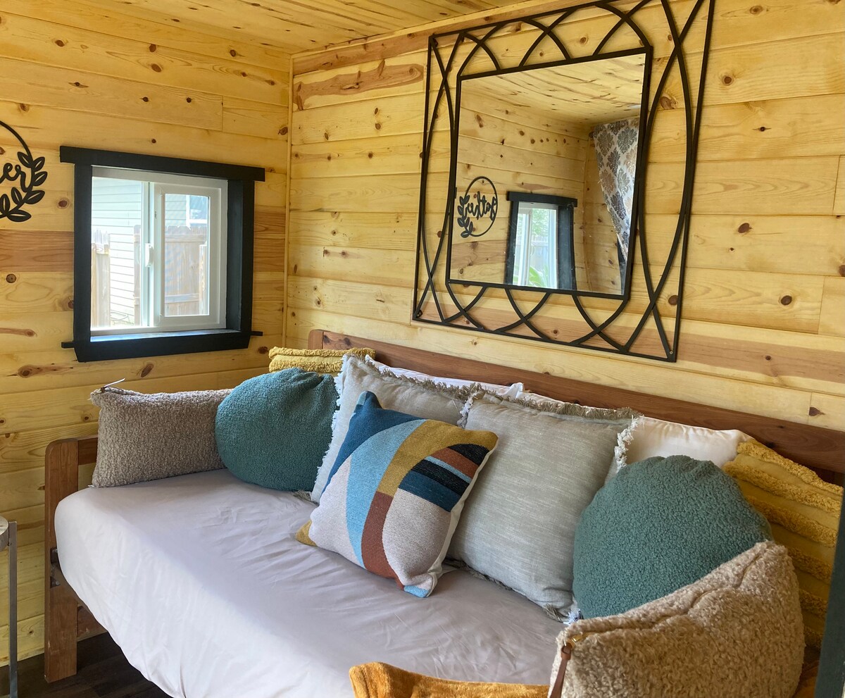 A cozy living area features a twin sofa bed layered with decorative pillows, providing a comfortable spot for relaxation. Light wood paneling lines the walls, while a mirror enhances the sense of space. A window offers a view of the fenced yard.