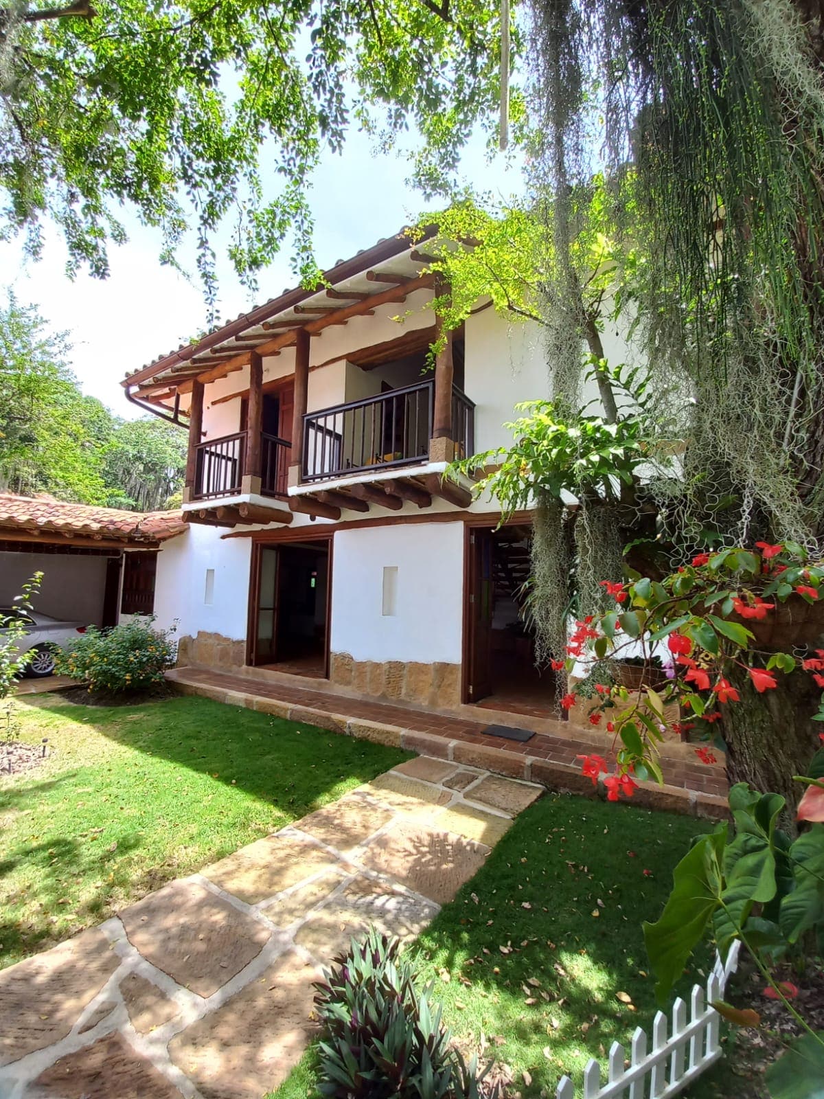 A two-story house is framed by lush greenery and flowers, featuring a stone pathway leading to the entrance. Balconies are visible on the upper floor, providing a view of the surrounding natural landscape. The exterior combines white walls with wooden beams.