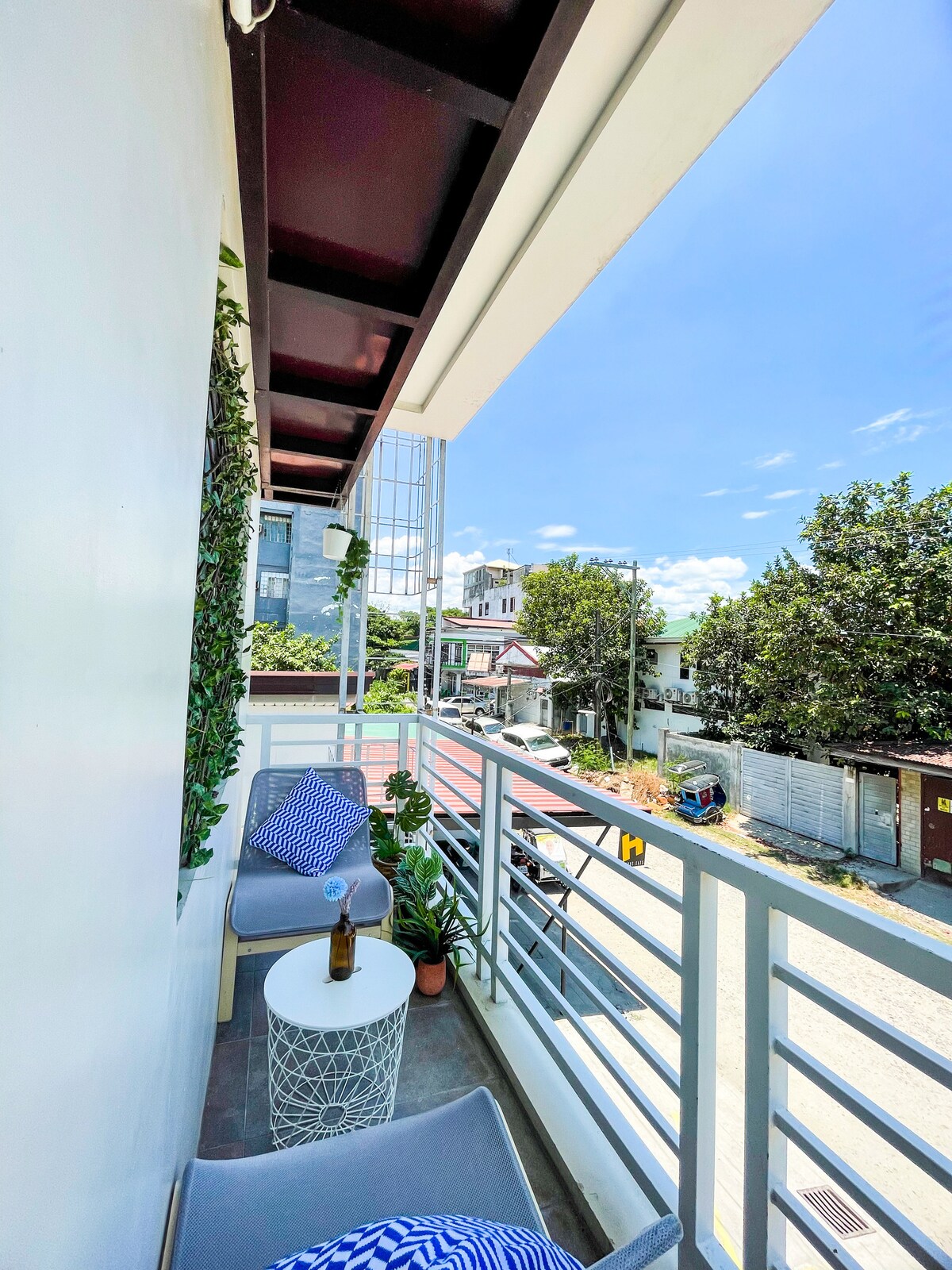 A balcony space is displayed with two comfortable chairs and a small round table. Lush green plants are positioned nearby, and a bottle is placed on the table. Bright sunlight illuminates the outdoor area, providing a view of the surrounding neighborhood.