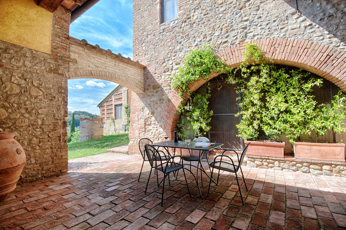 An outdoor terrace is highlighted by a dining table surrounded by metal chairs, set against a backdrop of stone architecture. Lush greenery cascades along the walls, and a scenic view of the countryside is visible through an archway, creating an inviting space for relaxation.