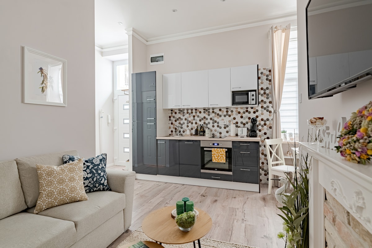 The modern kitchen is equipped with a range of appliances and features a sleek design with a blend of light and dark cabinetry. A comfortable seating area is highlighted by a light-colored sofa adorned with patterned cushions. A round coffee table sits on the textured rug, creating a cozy atmosphere.