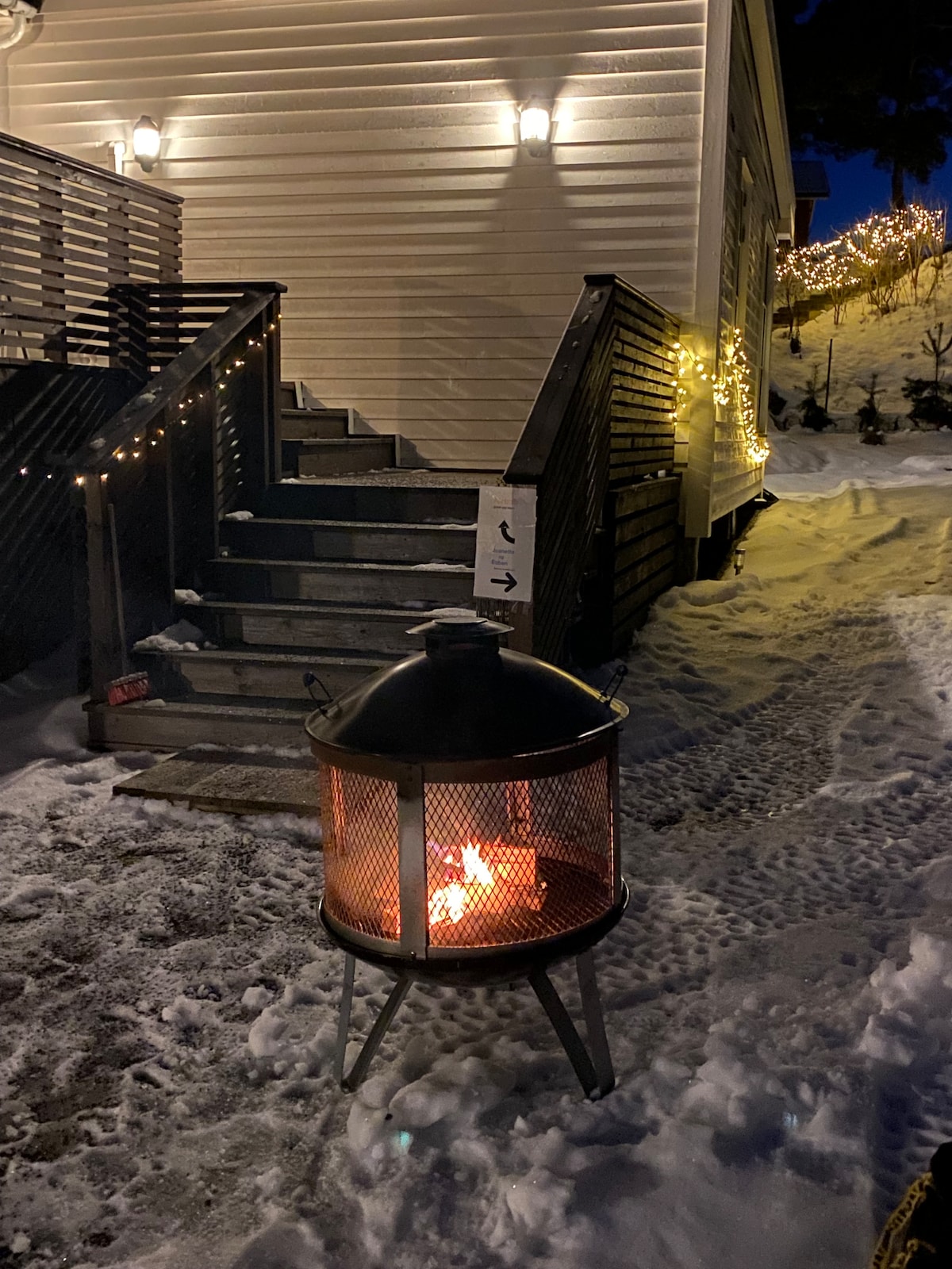 A warm glow from a fire pit is visible in the foreground, surrounded by snow. Steps leading to a house are lined with lights, providing a soft illumination against the winter landscape. The exterior of the house appears quaint and inviting, blending harmoniously with the surroundings.