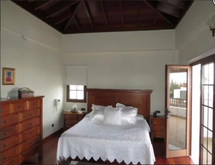 Chambre principale avec plafonds en bois surélevés, salle de bain attenante et doubles portes patio donnant sur le grand balcon