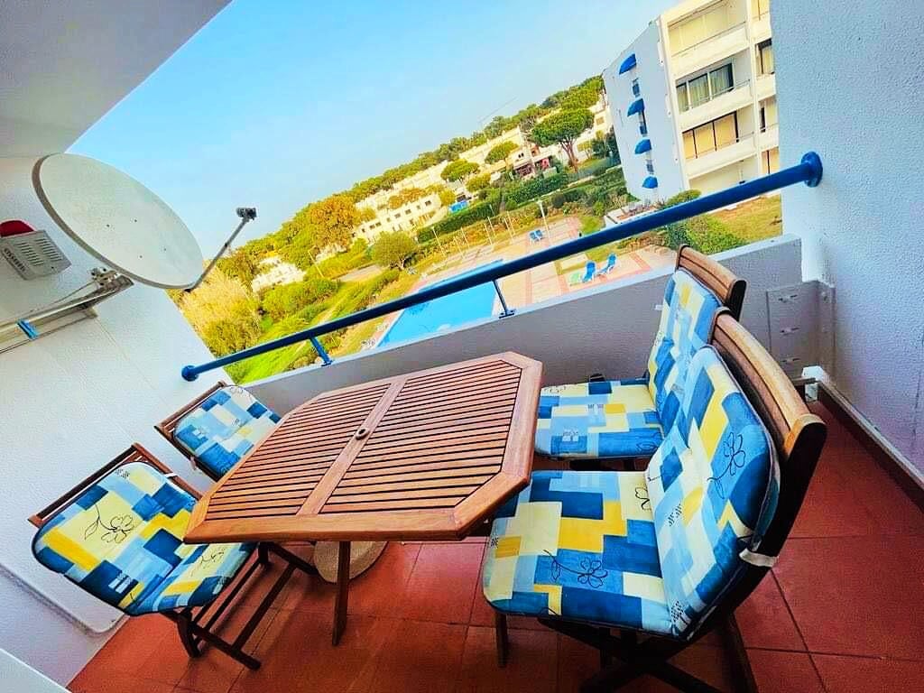 A balcony area is equipped with a wooden table surrounded by four cushioned chairs. The view overlooks a green landscape and two shimmering swimming pools, enhancing the outdoor setting. Bright blue skies are visible, complementing the relaxing atmosphere.