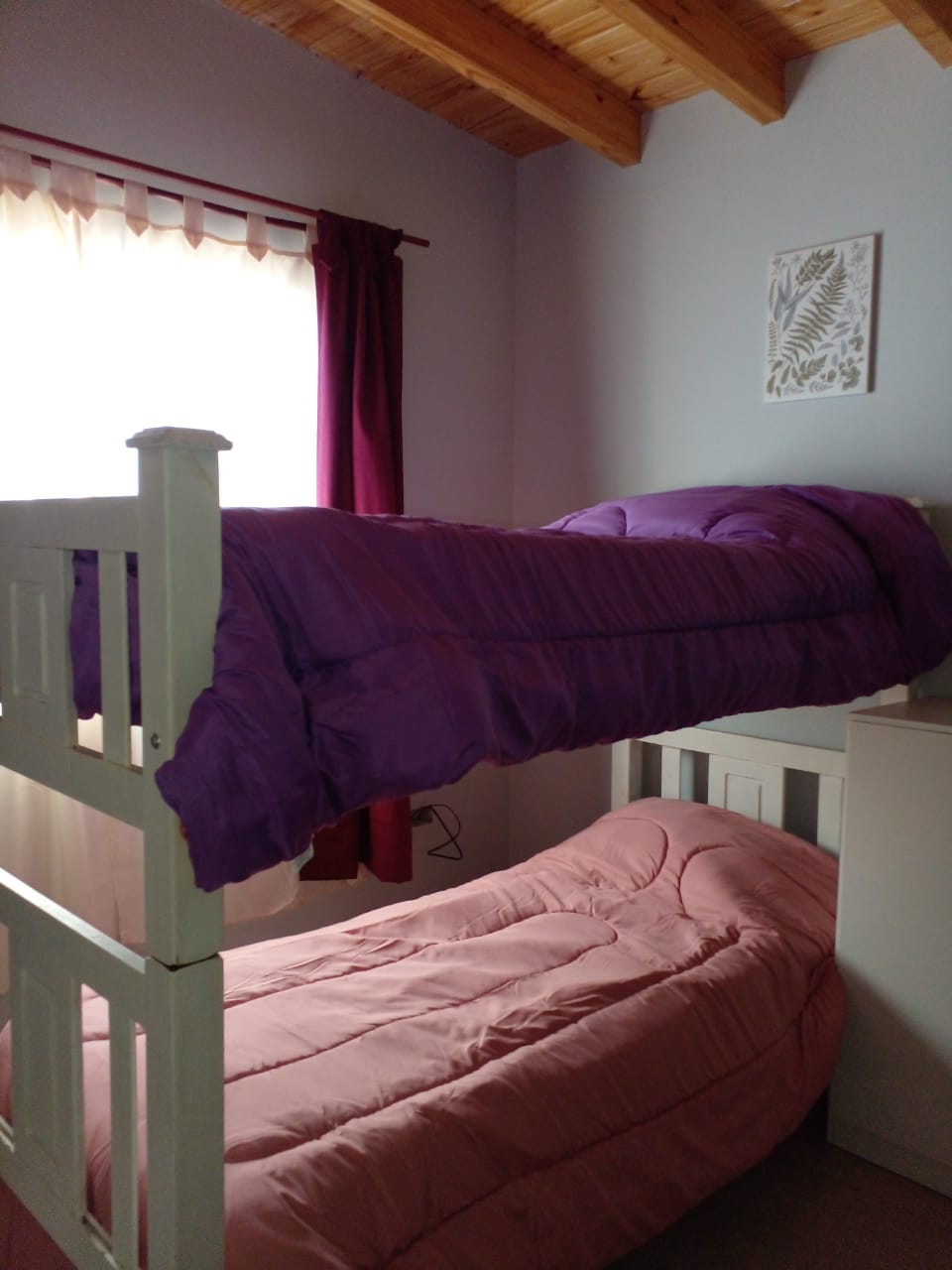 A bunk bed configuration occupies the corner of the room, featuring a purple comforter on the upper level and a pink comforter on the lower. Natural light filters through sheer curtains, softly illuminating the space, while a small decorative wall piece adds a touch of character.