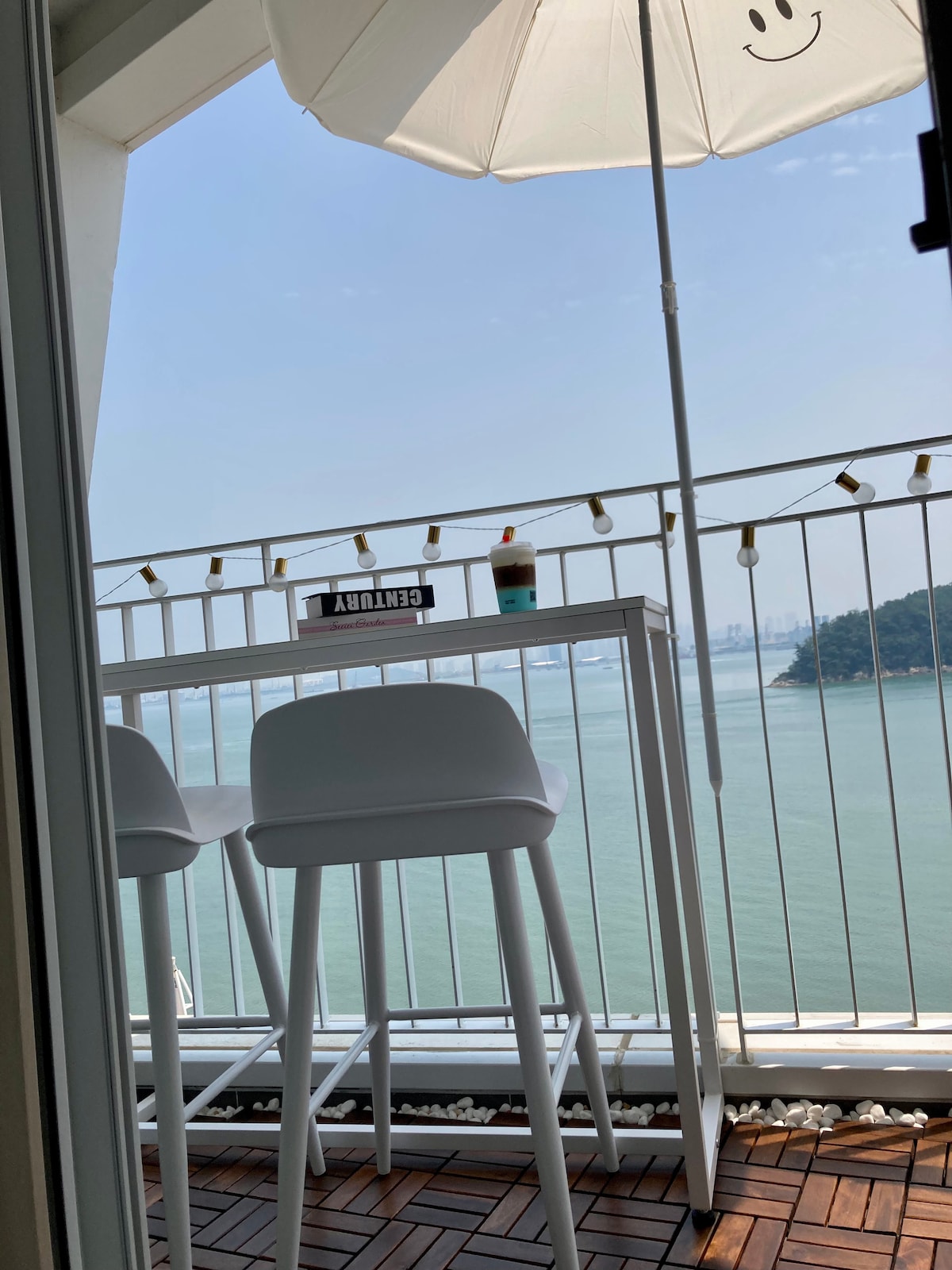 A modern white bar table with two high stools is positioned on a balcony overlooking the water. A sun umbrella provides shade, and string lights are arranged along the railing, creating a relaxing outdoor space with a scenic view.