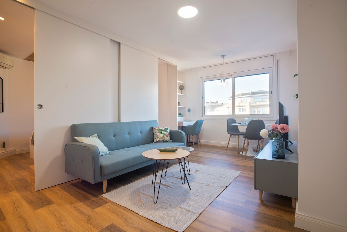 A bright living area features a gray sofa with decorative pillows, a round coffee table, and a rug on the wooden floor. A window offers natural light, and a dining area with chairs is visible in the background. A workspace with a desk is also present.