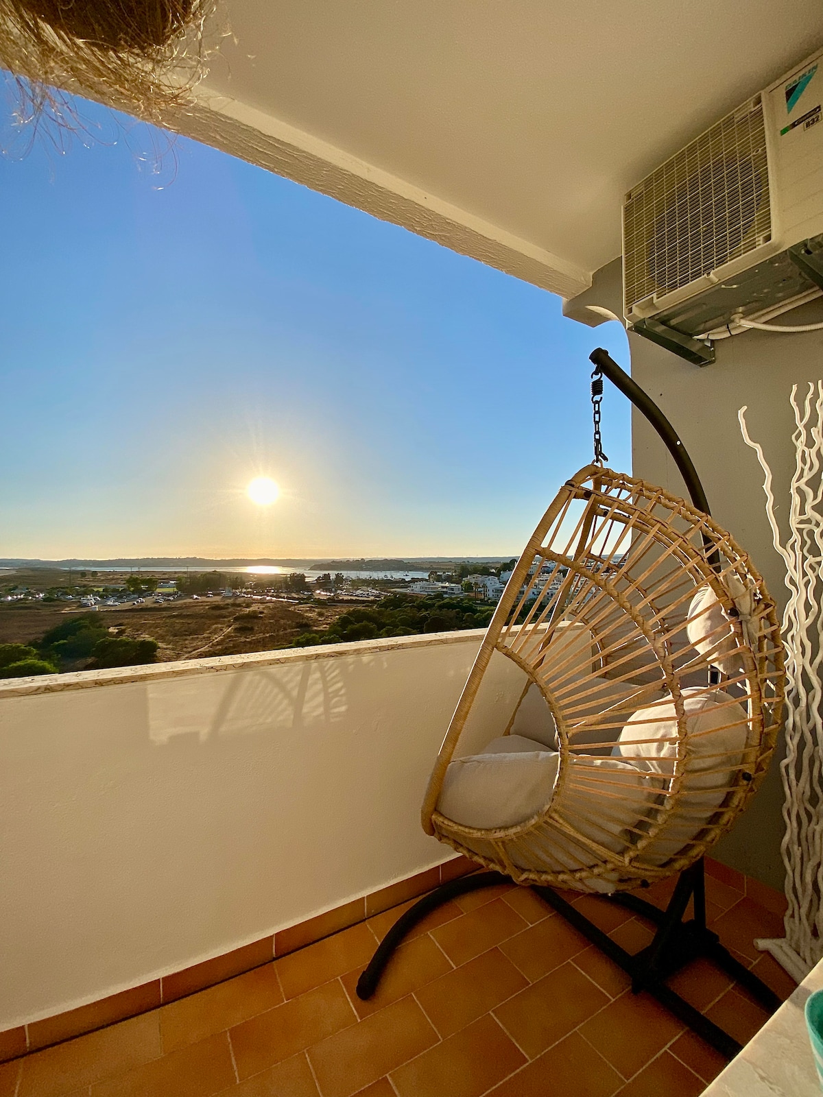 A hanging chair made of a woven material is positioned on a balcony, providing a scenic view of the sunset over the water. The soft glow of the sun enhances the calm atmosphere, while an air conditioning unit is mounted on the wall nearby.
