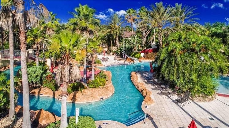 A resort-style pool area is surrounded by lush palm trees and vibrant greenery. A lazy river meanders through the space, leading to a water slide. Lounge chairs are placed throughout the area, inviting relaxation under the clear blue sky.