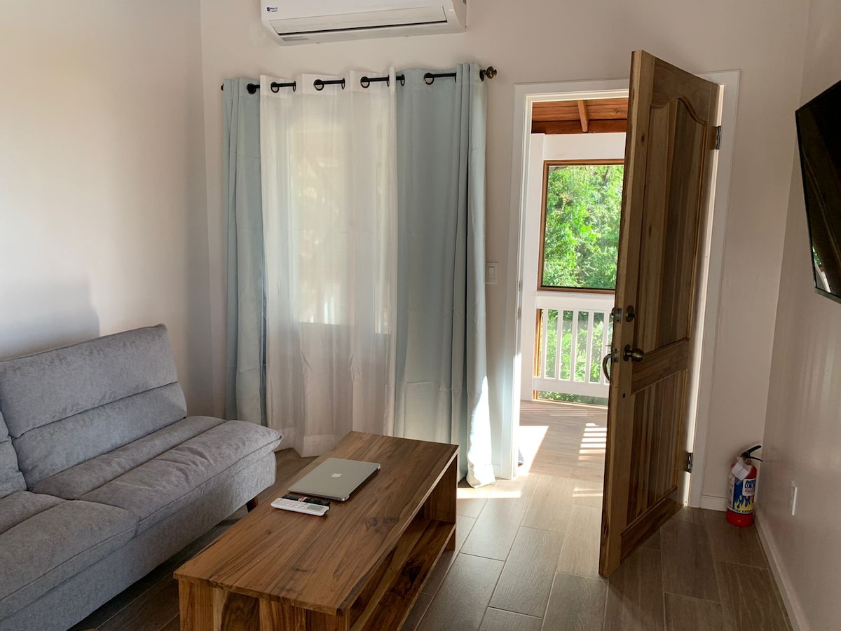 A cozy living area features a gray sofa and a wooden coffee table placed on tiled flooring. Light filters through sheer curtains, illuminating the space. An open door leads to a balcony surrounded by greenery, and a wall-mounted television is visible in the corner.