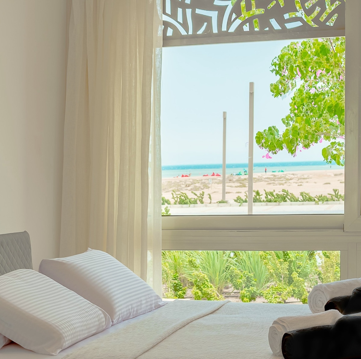 A spacious bedroom is provided with soft bedding and positioned near large windows. Sheer curtains allow gentle light to filter in, offering a view of the vibrant landscape and the azure sea beyond, framed by lush greenery.