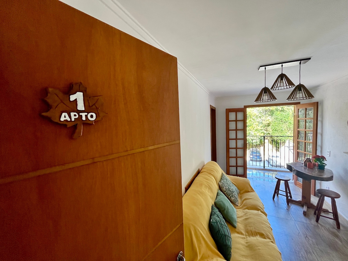 An apartment door is seen ajar, revealing a cozy living area with a large sofa adorned with green cushions. Natural light enters through French doors opening to a balcony, where a small table is set with a plant, adding warmth to the scene.