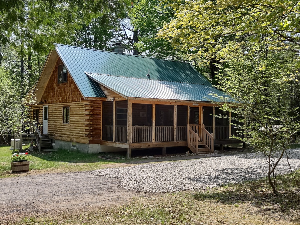 A charming log cabin with a metal roof is seen nestled among lush greenery. The front porch, screened for comfort, offers easy access from the gravel driveway. Nearby, decorative planters add a touch of nature to the welcoming entrance.