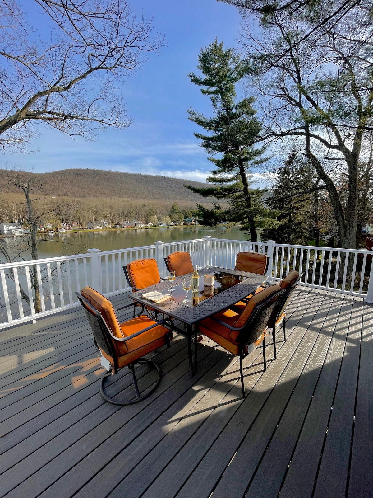 An outdoor dining set is positioned on a spacious deck, featuring a rectangular table surrounded by six cushioned chairs. The deck overlooks a serene lake and wooded hills, with clear skies above and trees providing shade.