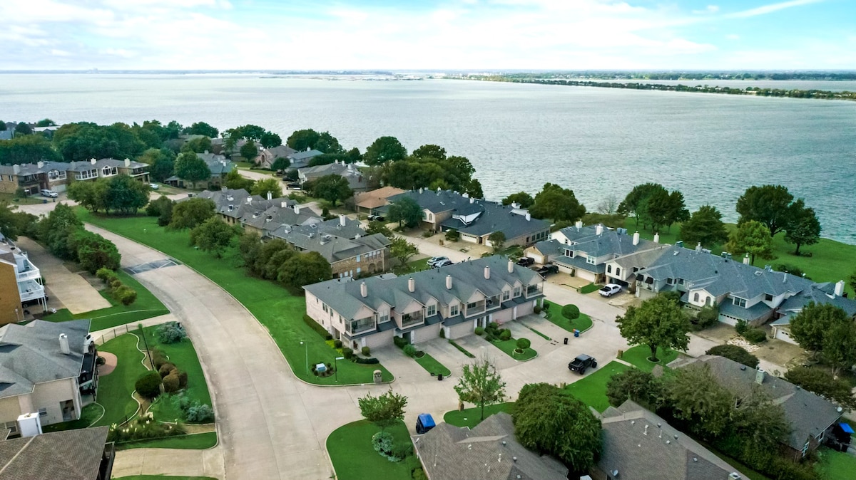 An aerial view captures the serene lakeside community, showcasing a grid of townhouses bordered by lush green lawns and mature trees. The expansive lake extends into the distance, with clear skies above, while residential driveways and winding roads are visible throughout the area.