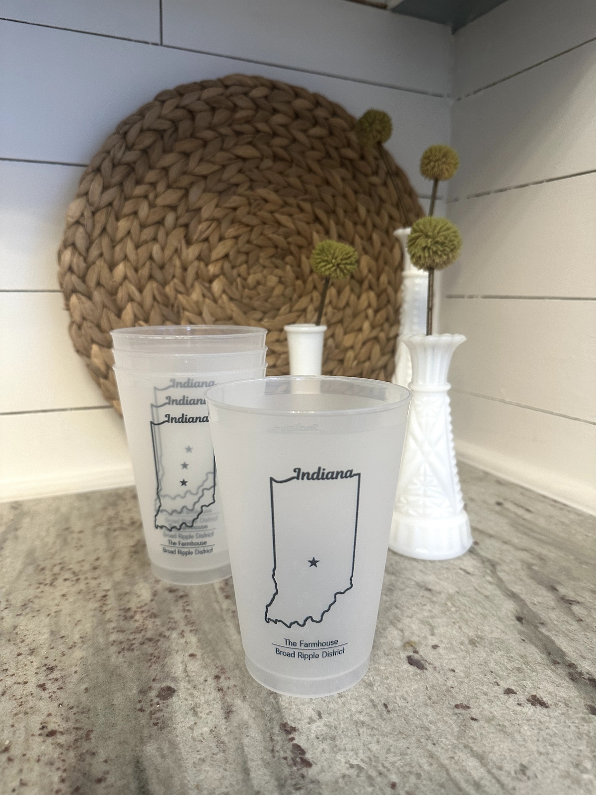 Frosted cups featuring an outline of Indiana with a star marking Broad Ripple District are displayed on a polished countertop. A woven placemat rests behind the cups, complemented by a simple floral vase holding green stems.