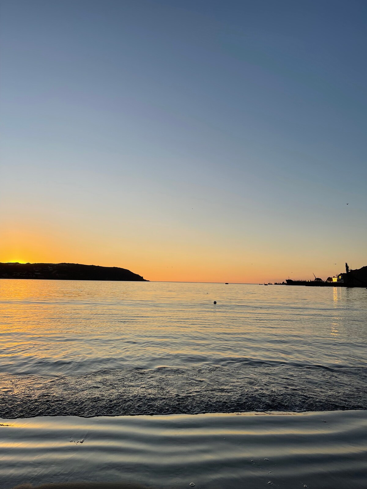 A serene view of the horizon at sunset, where warm orange and soft yellow hues blend into the calm waters. The silhouette of a distant hill is seen on the left, while gentle waves reflect the fading light. The tranquil atmosphere evokes a peaceful ambiance.