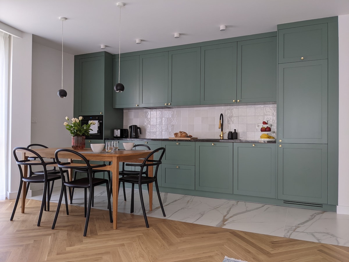 The kitchen features olive green cabinetry and a sleek countertop. A wooden dining table is surrounded by four black chairs. A variety of kitchen appliances are visible, including a coffee maker and kettle. Fresh flowers are displayed on the table, adding a touch of nature.