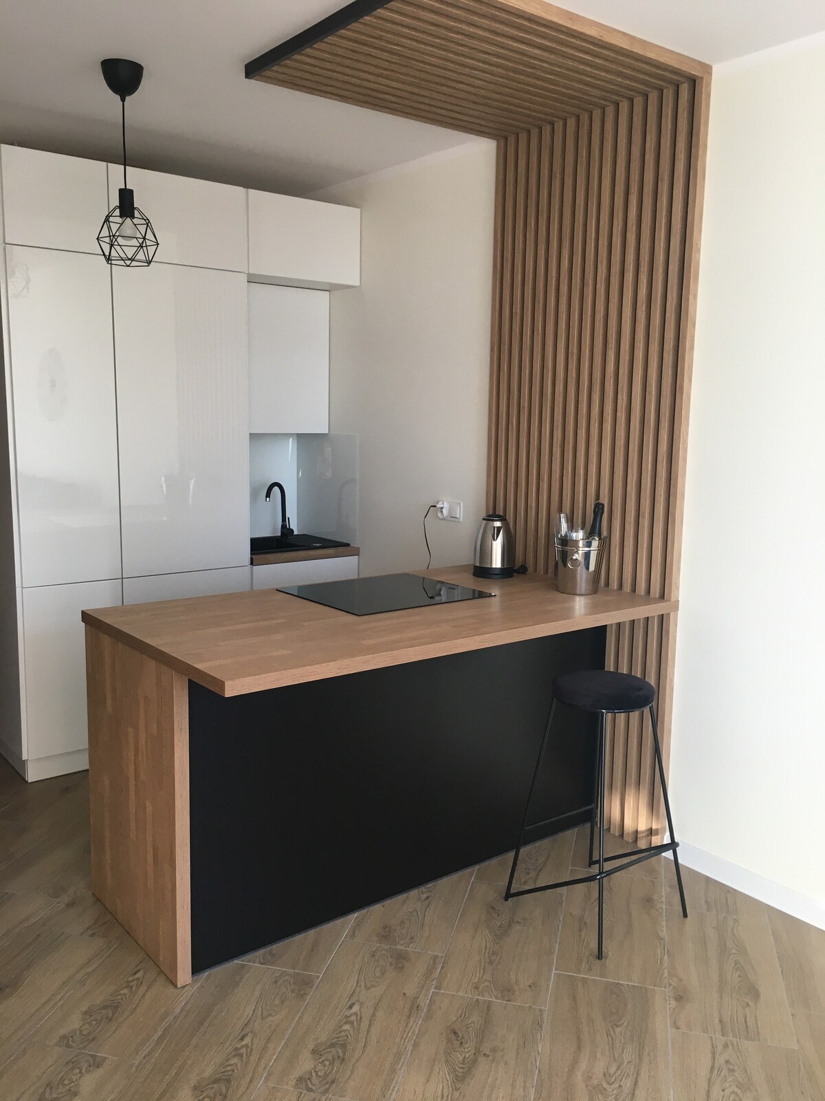 A modern kitchen area features sleek cabinetry with a combination of black and natural wood finishes. A compact wooden countertop provides space for meal preparation, complemented by minimalist bar stool seating. A stylish pendant light hangs above, enhancing the contemporary design.