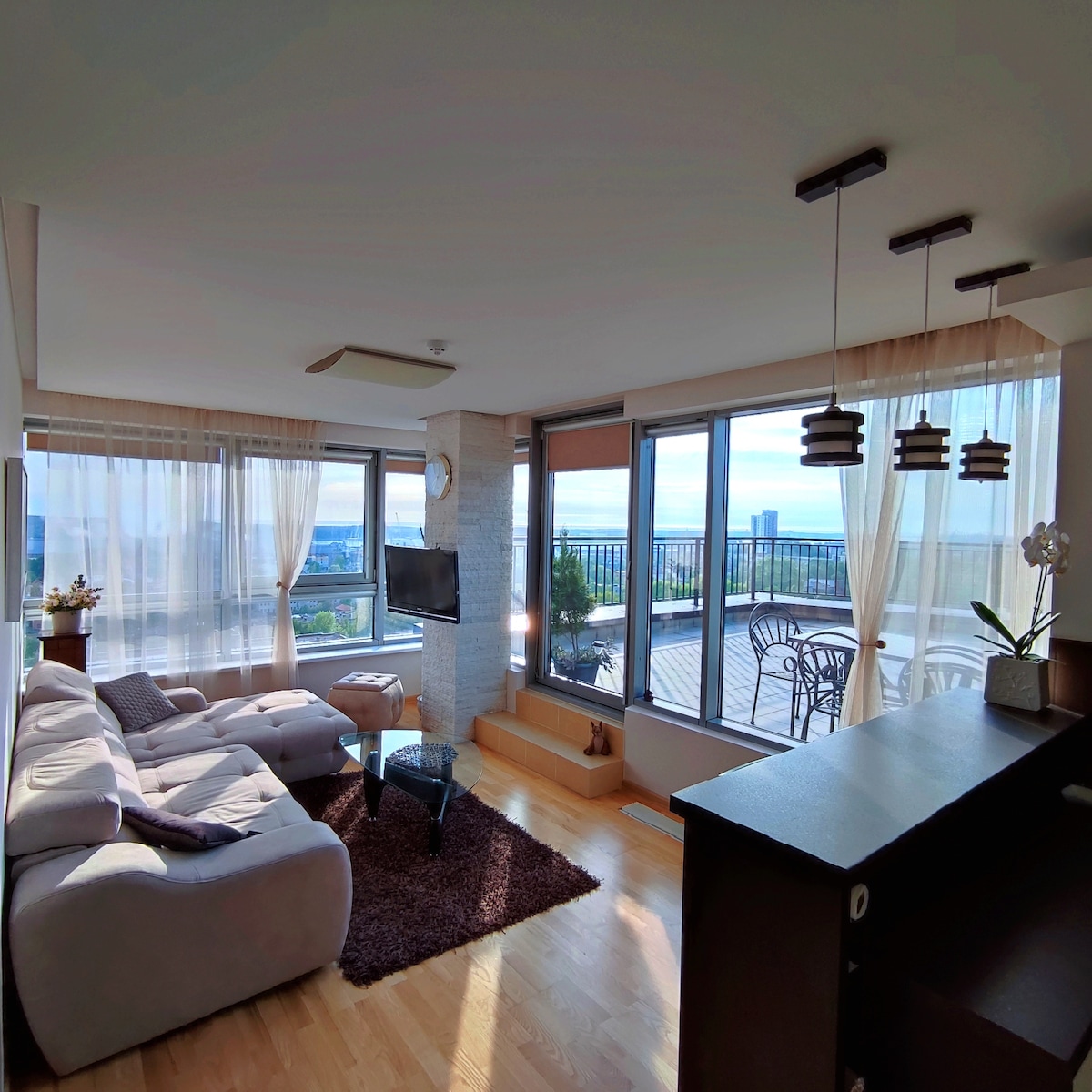 A bright living area features a spacious sectional sofa and a coffee table, complemented by a floor rug. Large windows provide views of the outdoor terrace and distant scenery, while soft curtains allow natural light to fill the space. Decorative plants add a touch of greenery.
