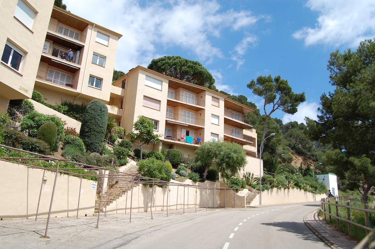 A residential building is situated on a hillside, featuring multiple levels with balconies and a landscaped garden. The smooth road runs alongside the property, highlighting the surrounding trees and greenery, while the architecture reflects a harmonious integration with the natural environment.