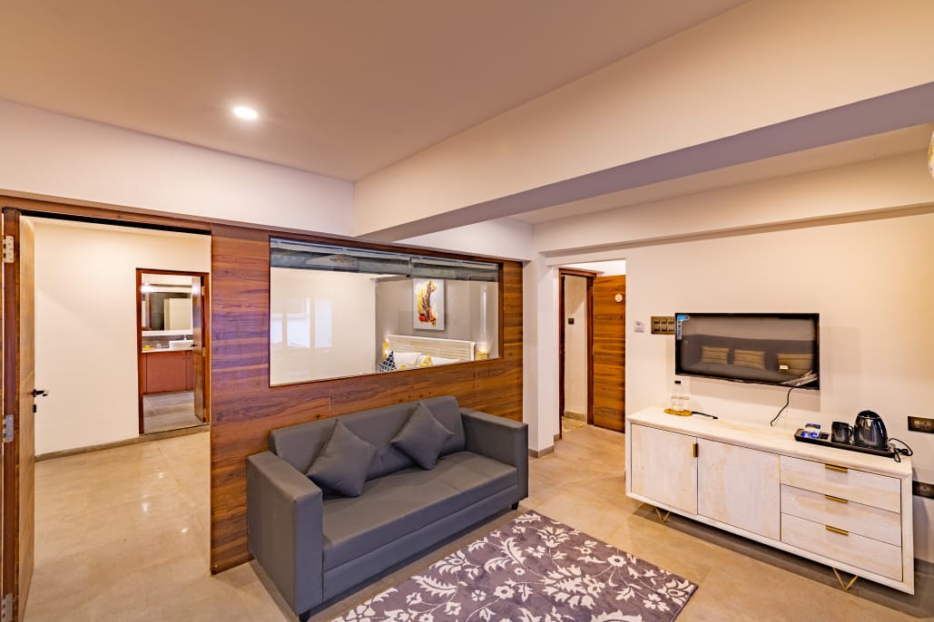 A spacious living area features a comfortable sofa and a modern TV console. Light walls are complemented by rich wooden accents, providing a warm ambiance. A partition offers a glimpse into the adjacent area, adding to the open feel of the layout.