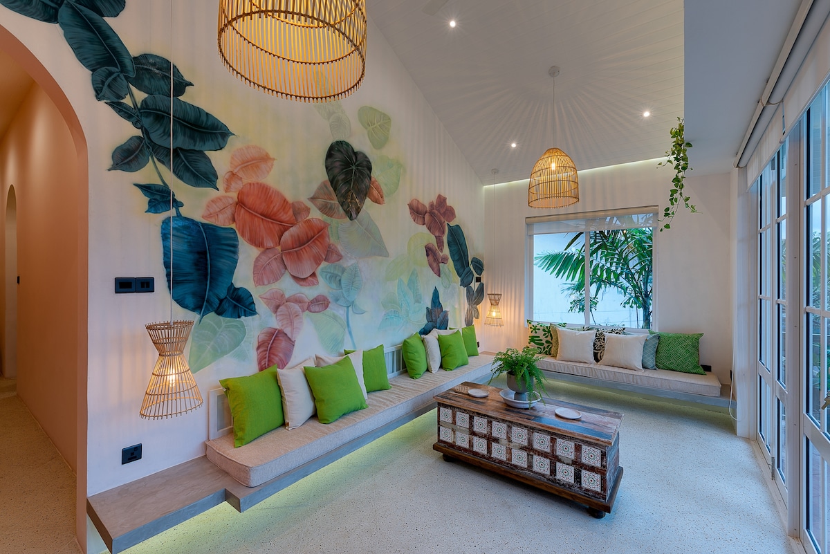 A modern living area features a large mural of tropical leaves in soft hues. Comfortable seating is arranged around a low wooden coffee table, with cushions in vibrant green shades. Natural light filters in through the adjacent window, accented by indoor plants.