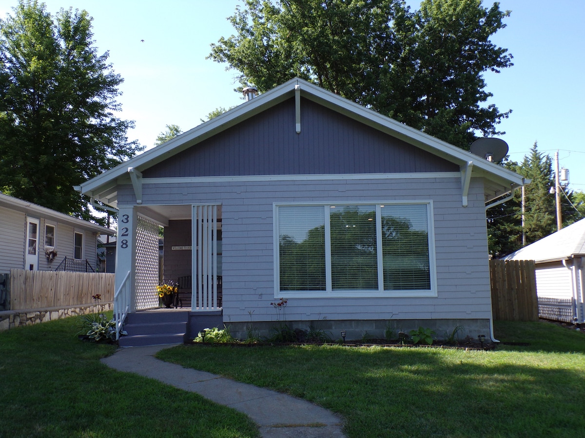 The exterior of a remodeled single-story home is shown, featuring a gray facade and a welcoming front porch. Large windows allow natural light to penetrate the interior, while a well-maintained lawn and paved walkway lead to the entrance.