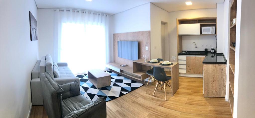 A modern living area is presented, featuring a light wooden floor with a geometric rug. A comfortable seating arrangement includes a gray sofa and two chairs. A dining table is set for two, and a wall-mounted television is visible. Natural light fills the space through large windows.