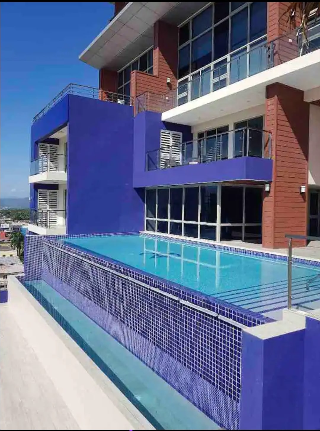An infinity pool extends along the edge of a modern building, framed by vibrant purple walls. Clear blue water reflects the sky and the surrounding skyline, enhancing the contemporary design of the condominium. A poolside area provides space for relaxation.