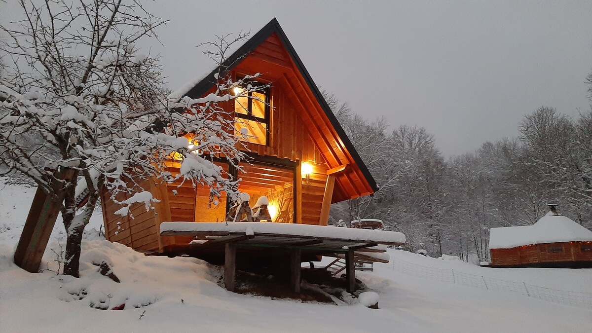A charming wooden chalet is seen surrounded by a snowy landscape, with warm lights glowing from within. The exterior features a spacious deck, while snow blankets the ground and branches of nearby trees, creating a serene winter atmosphere.