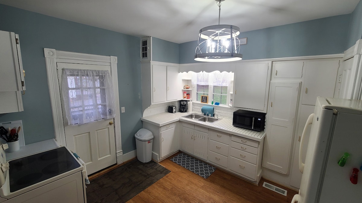 A fully equipped kitchen is visible, featuring white cabinetry and a light blue wall. Essential appliances include a full-size stove, refrigerator, microwave, and coffee maker. A window and door provide natural light, while a patterned rug lies on the wooden floor near the trash can.