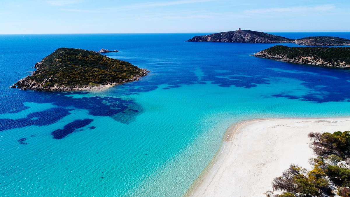 The image displays a vibrant coastal landscape, featuring clear turquoise waters and a sandy beach. Several small islands are visible in the distance, surrounded by greenery. The sky is bright and blue, creating a serene and inviting atmosphere.