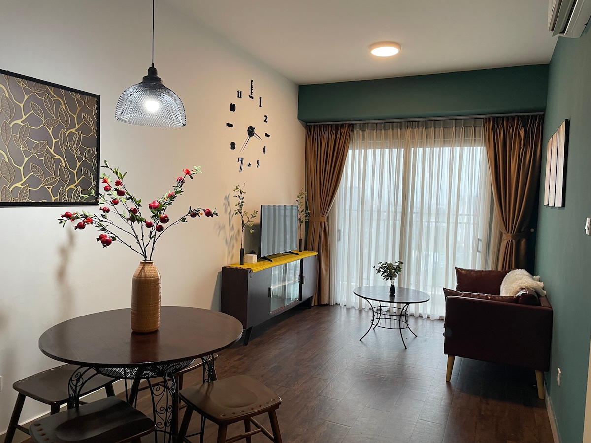 A modern living area features a round dining table surrounded by three sturdy stools. A decorative arrangement of flowers stands on the table. A comfortable armchair is positioned beside a flat-screen television on a stylish media console, with sheer curtains allowing natural light to filter in.