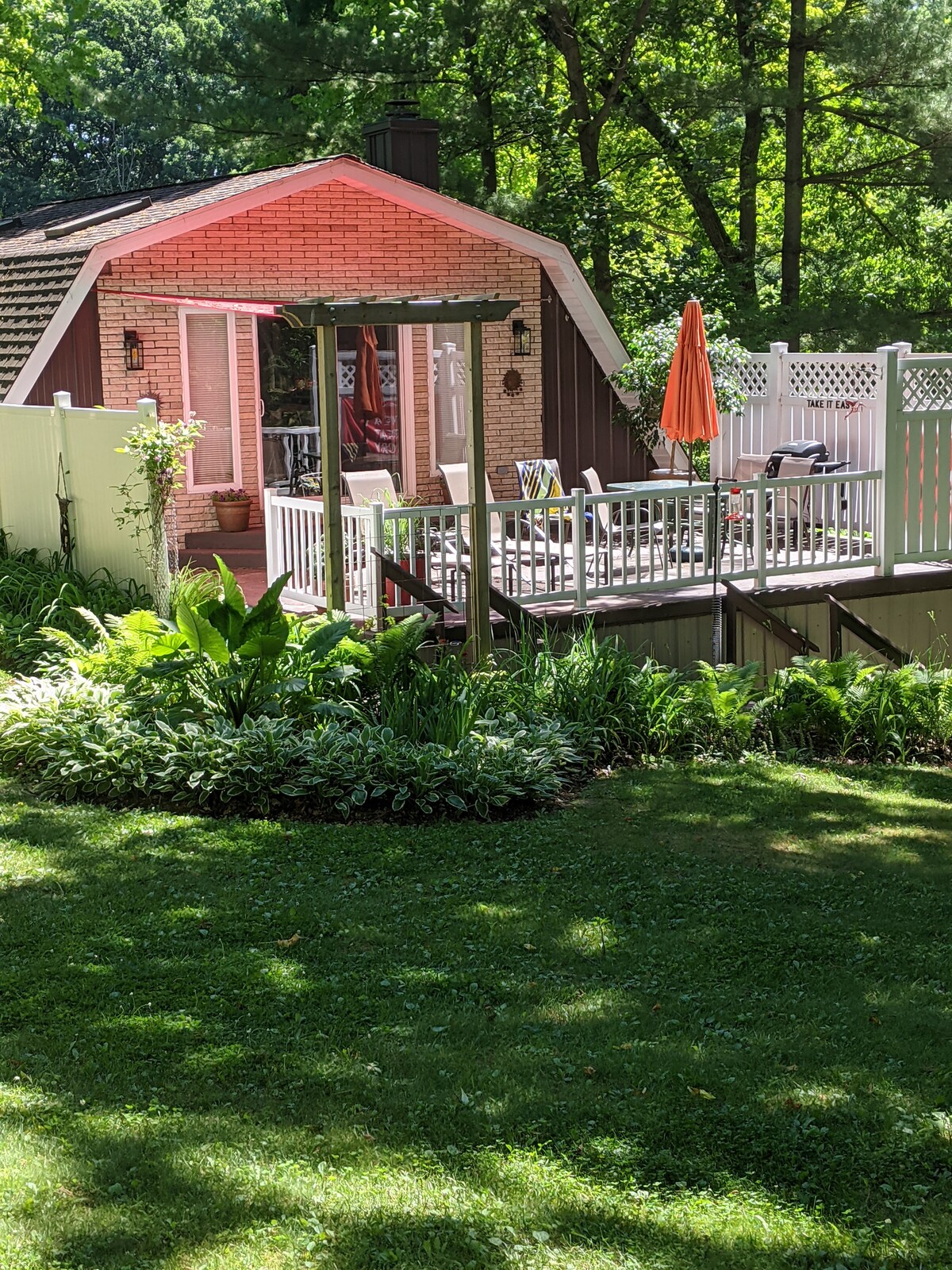 A well-maintained guest house is set amidst a lush green landscape. A large deck is visible, equipped with a gas grill and an umbrella-shaded table, surrounded by vibrant flowering plants. A white fence encloses the space, offering a sense of privacy.