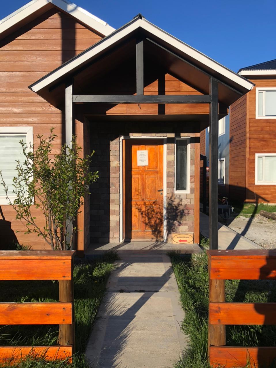 The entrance features a wooden front door framed by a light stone pathway. Natural greenery lines the walkway, and warm wooden fencing adds a rustic touch. A modern architectural design is observed in the angled roof and contrasting materials of the facade.