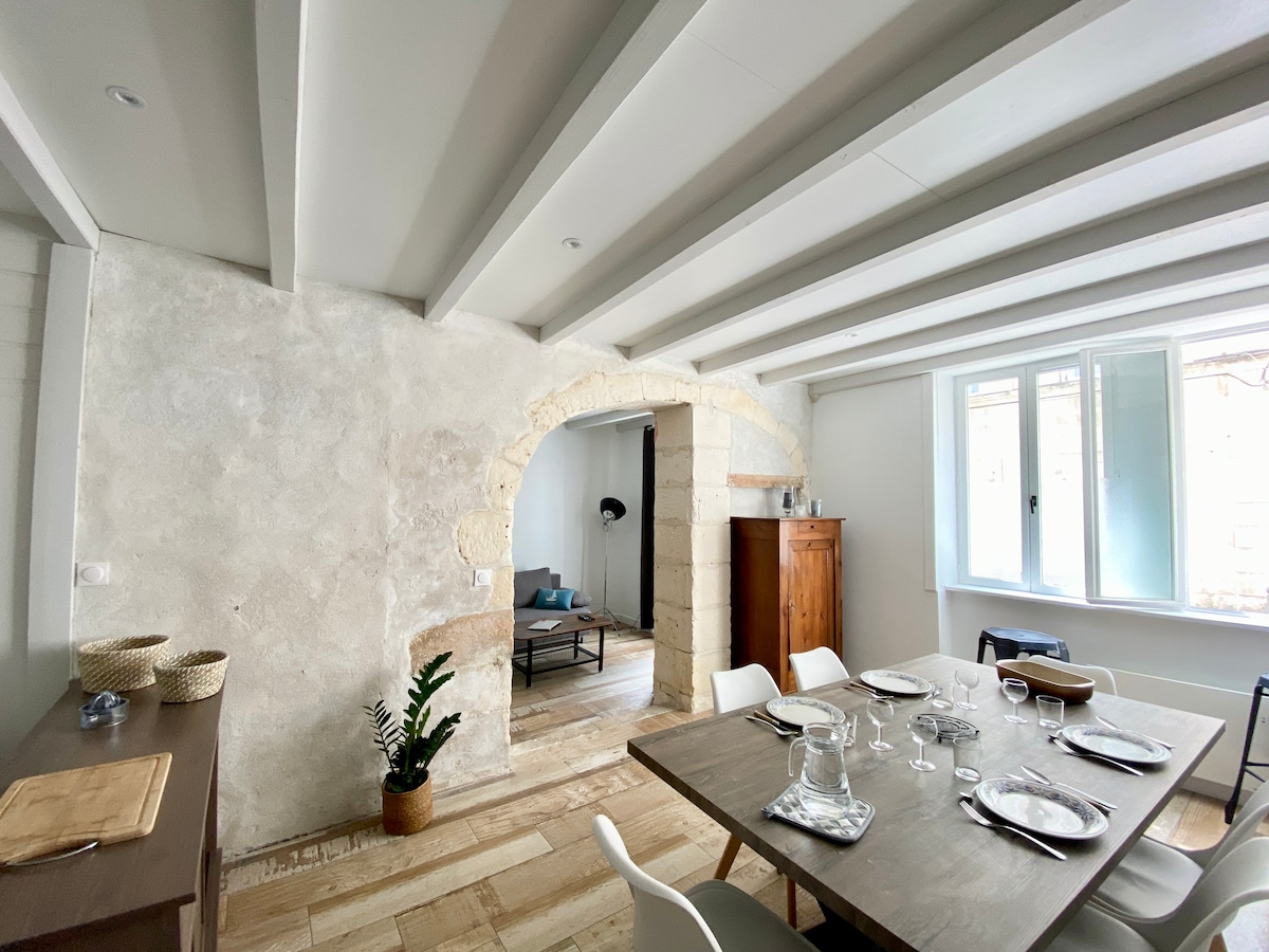 A dining area features a wooden table set with tableware, complemented by white chairs. An arched stone entryway leads to a living space with a couch. Natural light filters in through a window, highlighting the textured walls and ceiling beams.