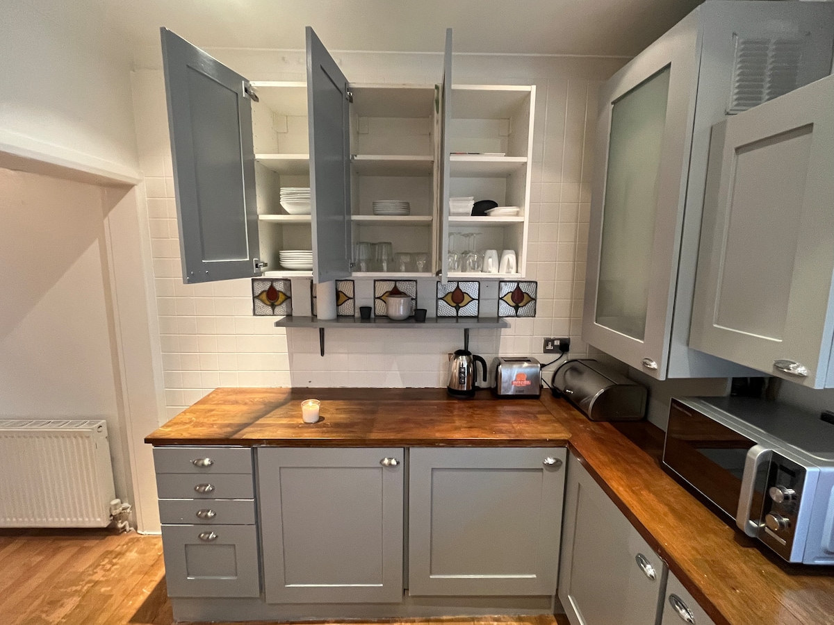 A functional kitchen space features gray cabinetry with open shelves displaying plates and glasses. A wooden countertop accommodates a kettle, toaster, and microwave. A candle rests on the surface, adding a subtle detail to the inviting atmosphere.