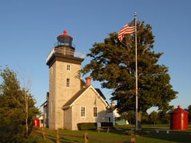 A historic lighthouse stands tall against a clear blue sky, highlighted by a red lantern atop the tower. An American flag waves gently in the breeze, and nearby structures include a stone building and red storage containers, set against a backdrop of lush greenery.