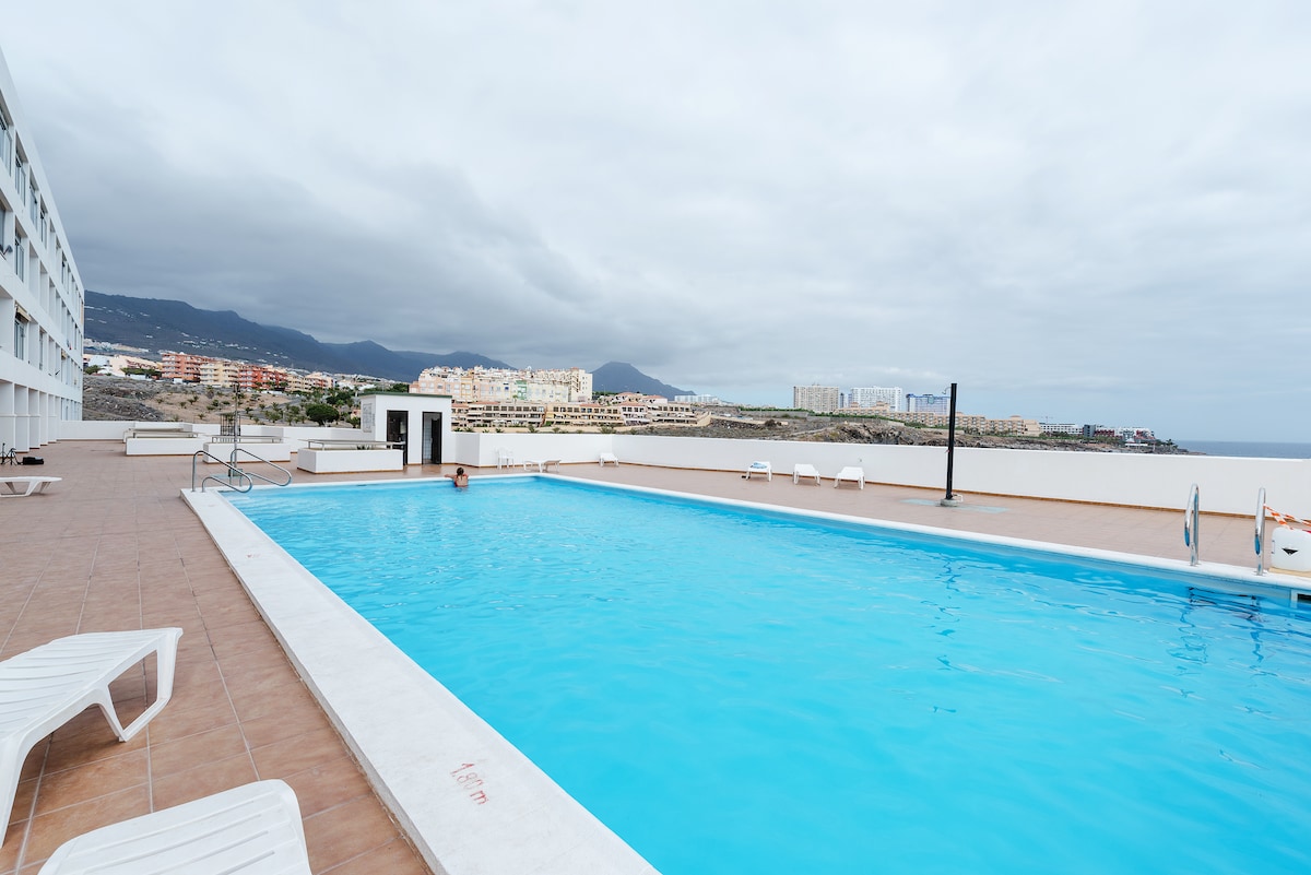 An outdoor swimming pool is visible, featuring clear blue water surrounded by a spacious deck. Several white lounge chairs are placed around the pool area. In the background, low buildings and hills can be seen under a cloudy sky.