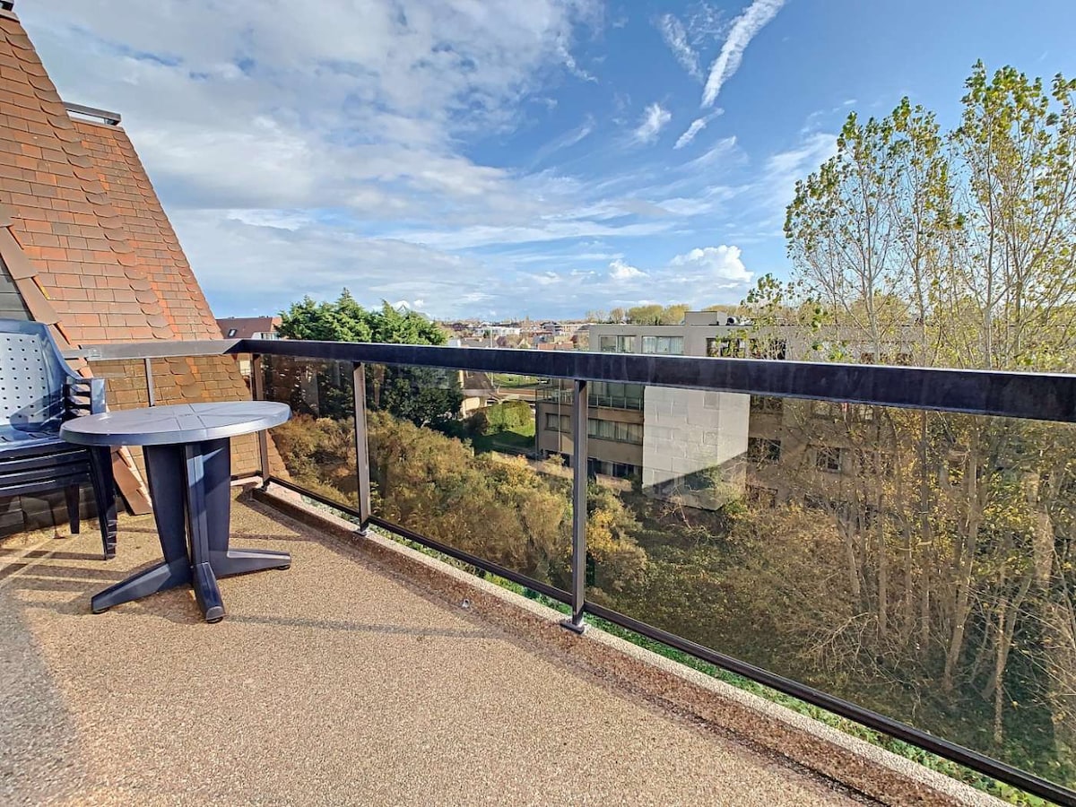 A balcony features a round table and chairs, overlooking a green landscape with trees and buildings in the distance. The sky is partly cloudy, allowing natural light to brighten the outdoor space.