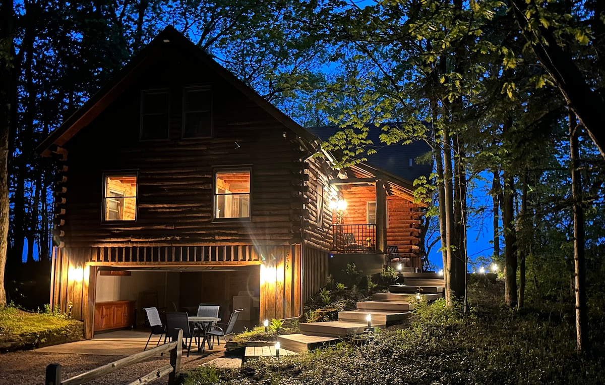 A cozy log cabin is illuminated at night, surrounded by trees. Soft lighting highlights the entrance and steps leading up to the covered porch. A seating area sits at the bottom of the steps, creating a welcoming ambiance amid the natural setting.