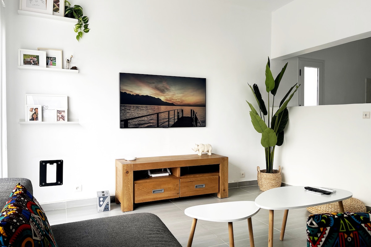 The living area features a modern design with a cozy sofa adorned with colorful cushions. A low wooden table sits in front of the sofa, complemented by an entertainment unit below an art piece depicting a serene landscape. Natural light is welcomed through a window beside a decorative plant.