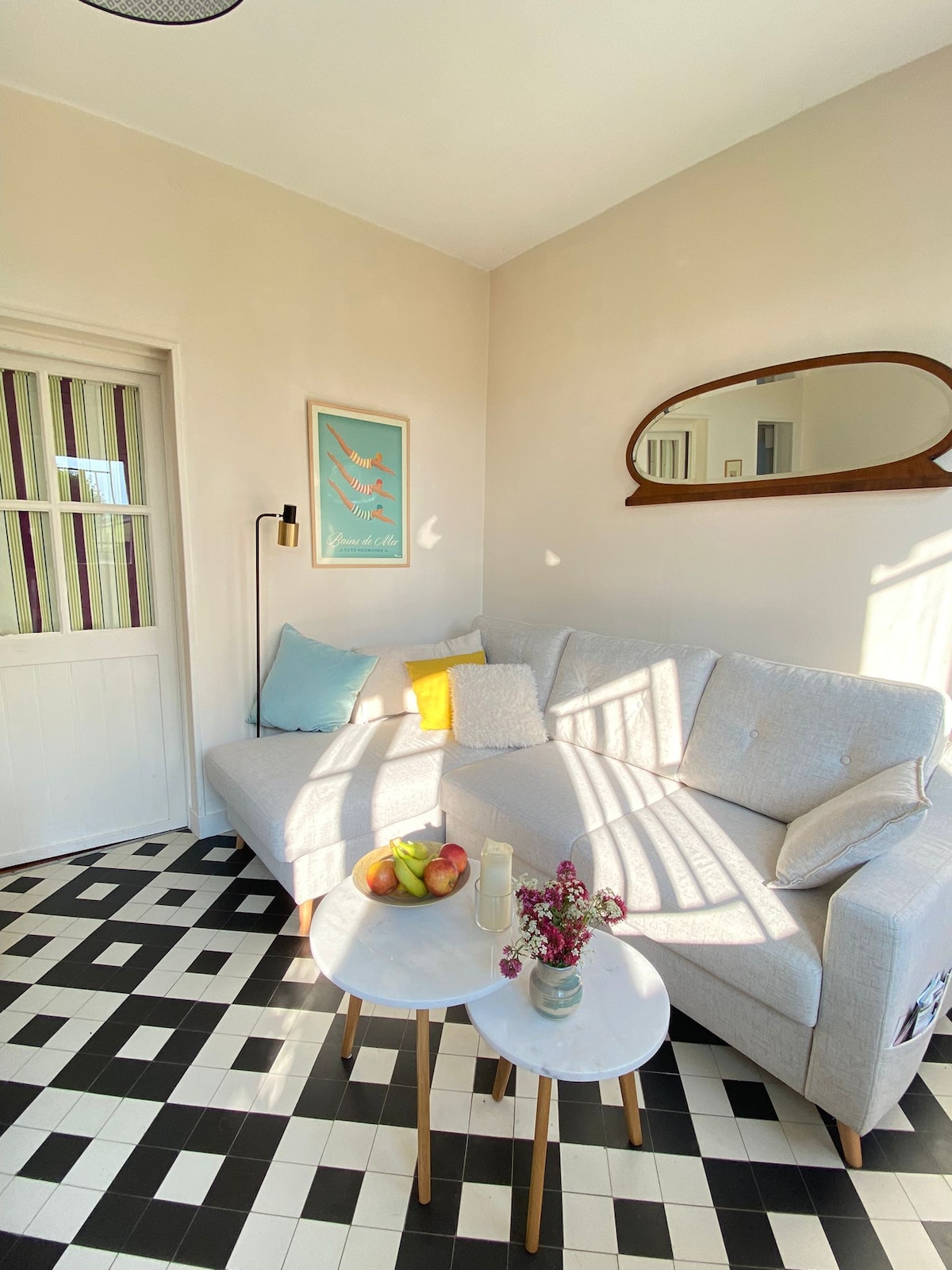 A light-filled living area features a gray sofa with pastel cushions, positioned next to a wall adorned with artwork. A pair of small tables support fresh fruit and a decorative vase, while patterned flooring adds an element of visual interest.