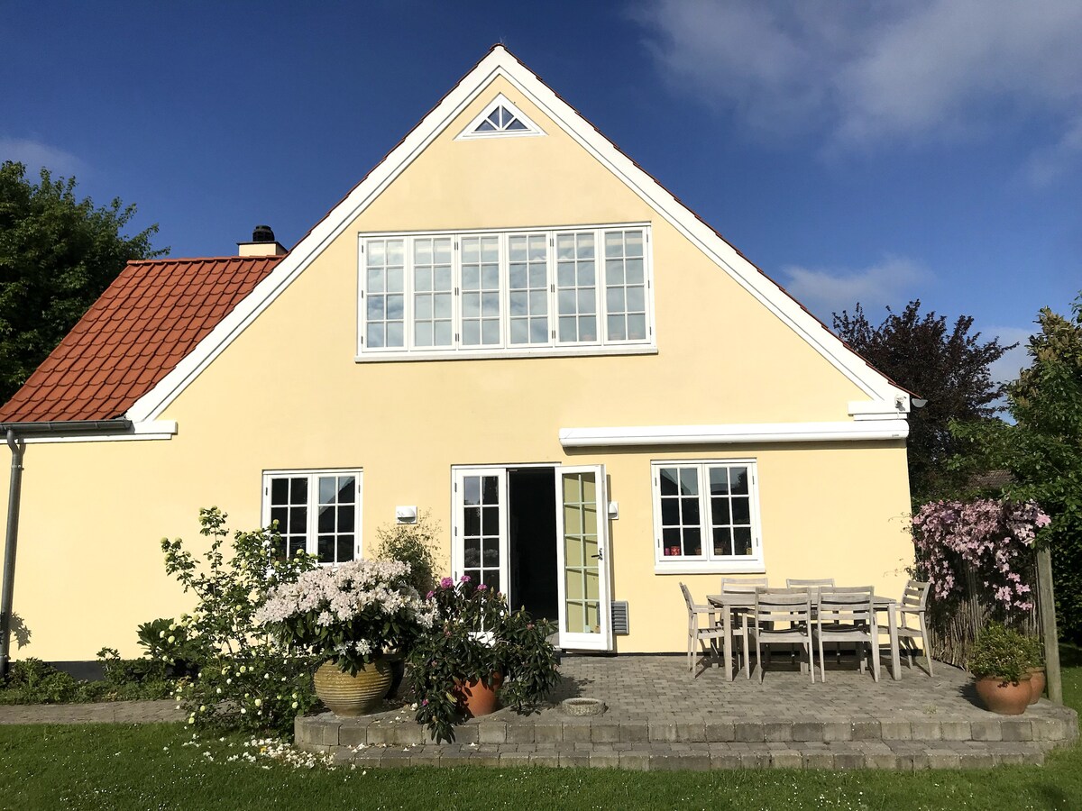 A charming yellow house is presented with a sloped roof and large front windows. The outdoor patio features a spacious dining table surrounded by flower pots, creating an inviting space for relaxation. The surrounding greenery enhances the house's welcoming appearance.