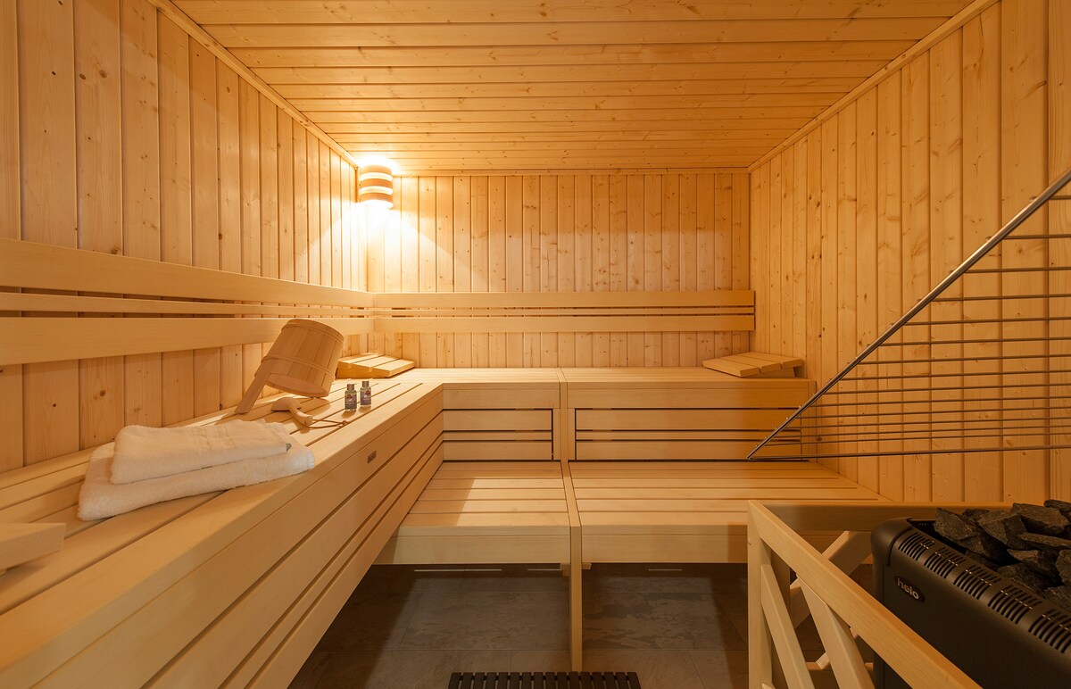 The sauna features wooden benches arranged in a spacious layout, complemented by warm lighting on the wooden walls. A heated stone unit is present, and towels are neatly placed nearby, creating a serene environment for relaxation.