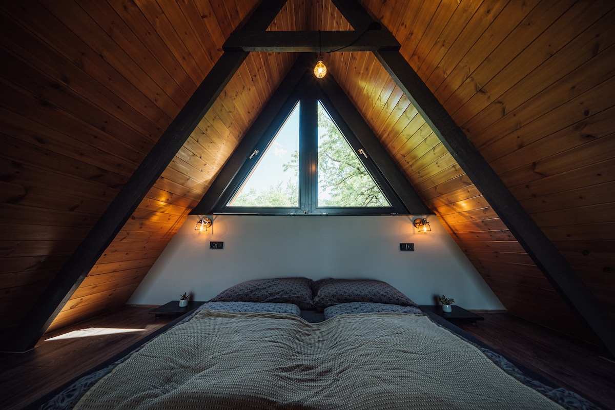 An A-frame bedroom features a spacious bed adorned with a neutral blanket. Natural light enters through large, triangular windows, which are complemented by accent lighting on either side. The warm wooden ceiling adds a cozy touch to the overall design.