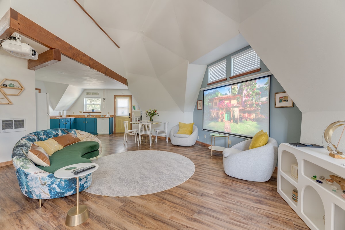A bright and airy living space showcases a large projection screen displaying colorful imagery. Soft furnishings include a rounded green sofa and two light-colored chairs, arranged around a circular area rug. Light wood floors contribute to the room's open feel, complemented by natural lighting from two windows.