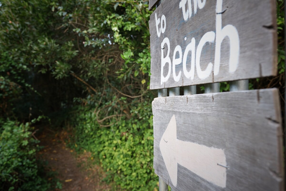 A weathered wooden sign directs towards the beach, featuring an arrow pointing left. Lush greenery surrounds the pathway leading to the beach, creating a natural frame for the route ahead.