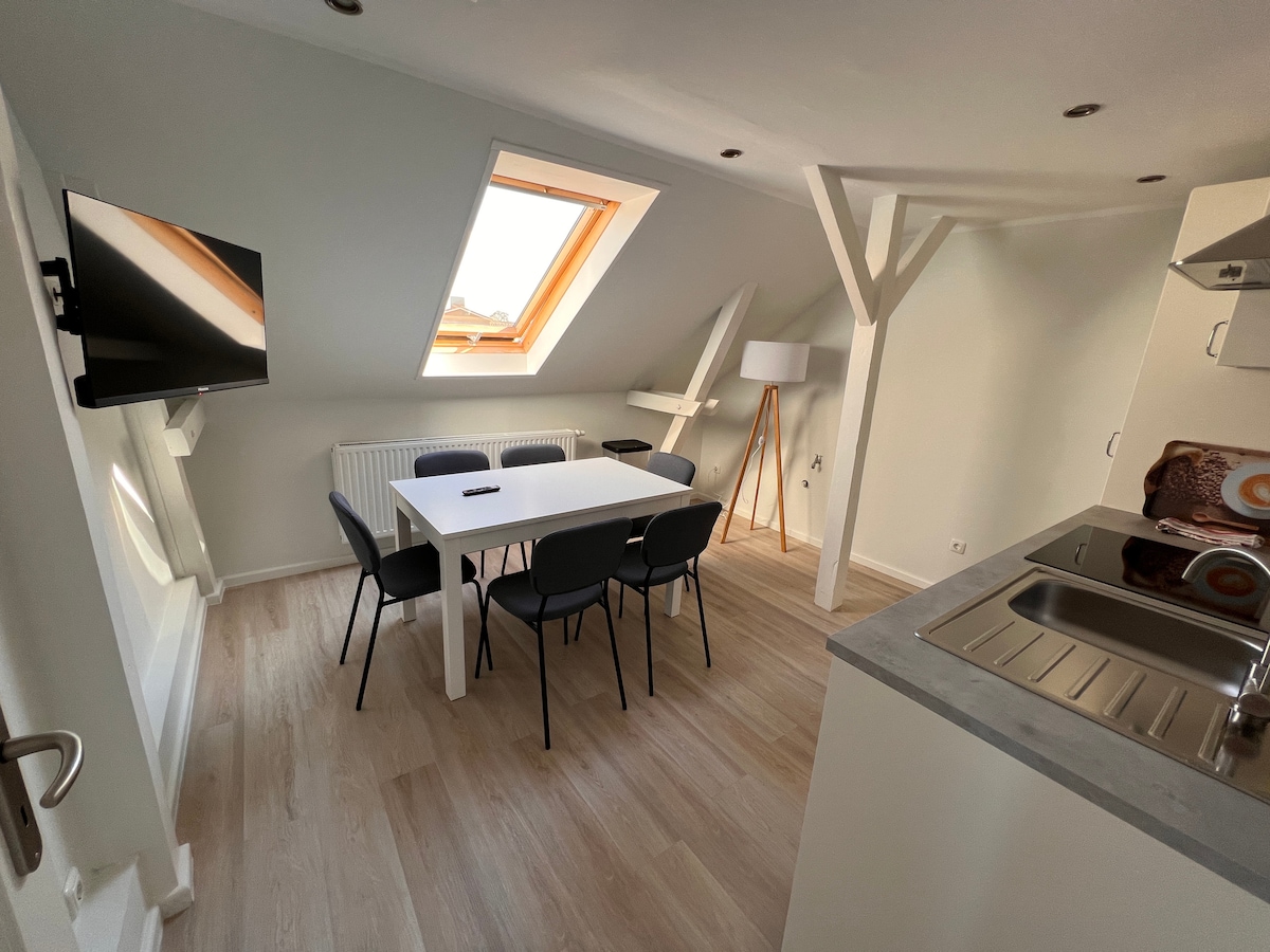 A dining area is presented with a white table surrounded by six black chairs. A television is mounted on the wall, and a sloped ceiling features a skylight allowing natural light. A kitchen island with a stainless-steel sink is visible, enhancing functionality.